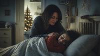 Adrianna sits on the edge of a bed in early morning light, waking her daughter.